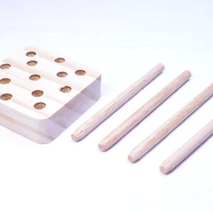 May include: A wooden peg game with a square base featuring multiple circular holes. Four wooden pegs are positioned to the right of the base. The game is made of light-colored wood.