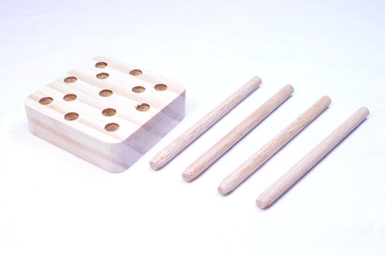 May include: A wooden peg game with a square base featuring multiple circular holes. Four wooden pegs are positioned to the right of the base. The game is made of light-colored wood.