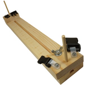 May include: Wooden arrow jig with two black plastic clamps and a wooden dowel. The jig has a groove for the arrow shaft and a ruler marked in inches.