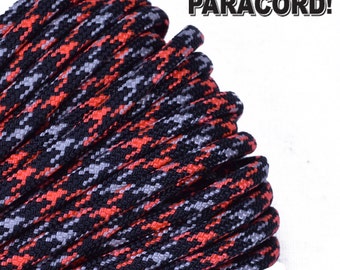Puede incluir: Primer plano de un paracord negro con un patrón de camuflaje rojo y gris. El paracord está enrollado y listo para ser usado para manualidades o supervivencia.