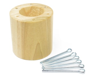 May include: A wooden dowel with multiple holes drilled into the top and side. A pile of silver cotter pins are in front of the dowel.