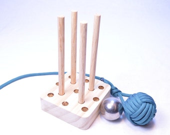 May include: A wooden peg game with four upright pegs, a silver ball, and a teal rope knot. The pegs are set in a light-colored wooden base. A teal rope is attached to the base and the knot.