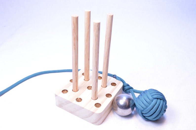 May include: A wooden peg game with four upright pegs, a silver ball, and a teal rope knot. The pegs are set in a light-colored wooden base. A teal rope is attached to the base and the knot.