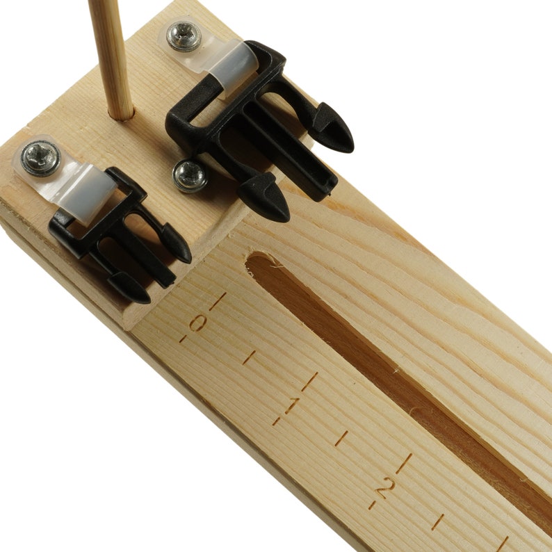 May include: A wooden jig with two black plastic buckles and a ruler marked from 0 to 2. The jig is used for making adjustable straps.