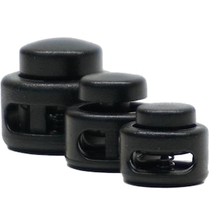 May include: Three black cord locks of varying sizes are arranged in a row. Each cord lock has a cylindrical body with a central hole and a spring mechanism. The top of each lock is a rounded button. The cord locks are made of a matte black plastic.