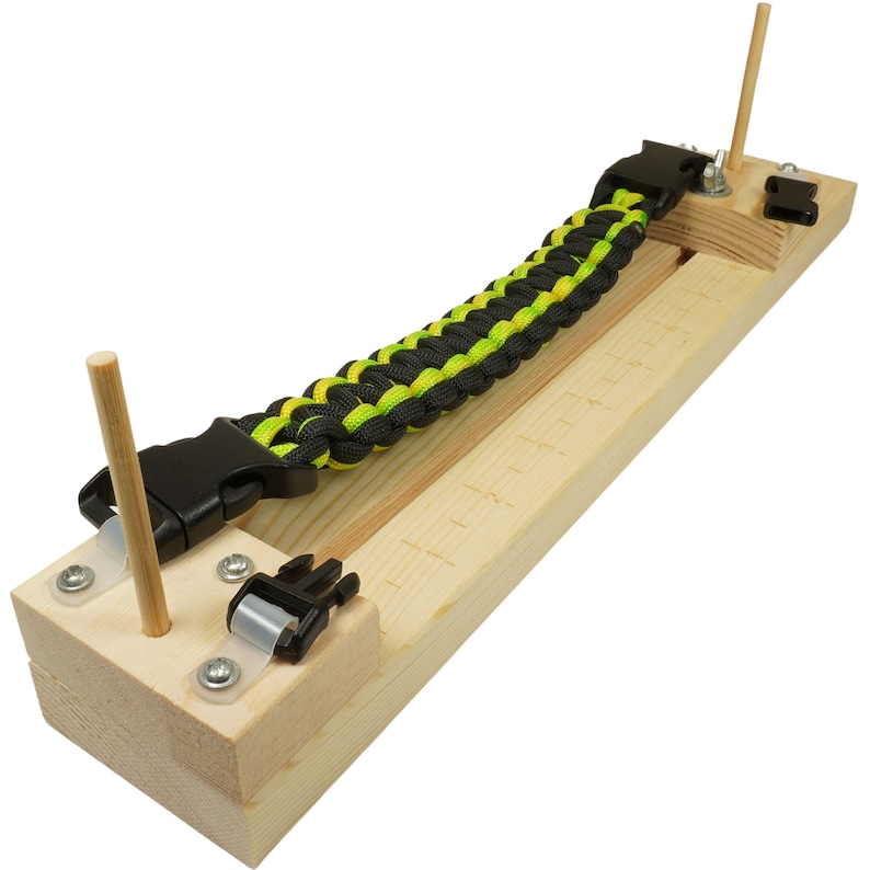 May include: A wooden paracord bracelet loom with a black and green paracord bracelet partially woven on it. The loom has two wooden dowels and two plastic buckles.