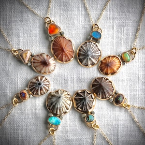 May include: A collection of handcrafted necklaces with gold chains. Each necklace features a unique pendant with a shell-like design, accented with colorful gemstones in shades of orange, blue, and green. The pendants are set in gold-colored metal.