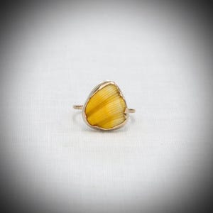 May include: A gold-colored ring featuring a unique, yellow, petal-shaped stone. The stone has a textured, striped pattern. The ring is set against a white background, highlighting the jewelry's design and color.