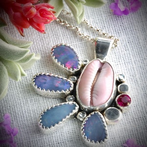 May include: A silver pendant necklace featuring a central pink seashell surrounded by blue-purple opals and a small red gemstone. The pendant is attached to a silver chain, showcasing a unique, nature-inspired design.