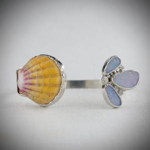May include: A silver ring featuring a seashell and a cluster of blue-toned opals. The seashell is yellow and orange with a pink interior. The opal cluster has a floral design. The ring is set against a white background.