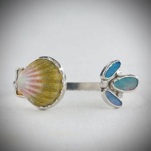 May include: A silver bracelet featuring a seashell and a cluster of blue opal stones. The seashell has yellow, pink, and white hues. The opal stones are oval and teardrop-shaped. The bracelet is on a white surface.