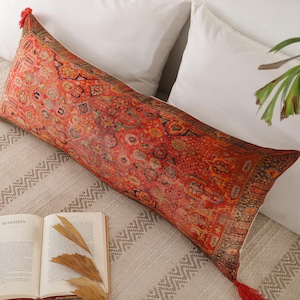 May include: A long, rectangular cushion with a vibrant, intricate pattern in shades of red, orange, and brown, reminiscent of a Persian rug. The cushion has red tassels at each end. The cushion is resting on a bed with white pillows and a patterned blanket.