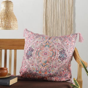 May include: A decorative square pillow with a detailed, colorful pattern in shades of pink, blue, and orange. The pillow has pink tassels on the corners and is placed on a wooden chair. The pillow's design resembles a traditional rug.