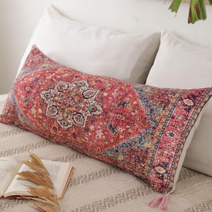 Puede incluir: Un cojín rectangular decorativo con un diseño de alfombra oriental roja, azul y crema. El cojín tiene borlas rosas en cada extremo y está colocado en una cama con almohadas blancas.