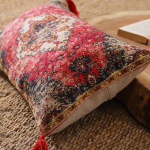 May include: Rectangular decorative pillow with a vibrant red, pink, and cream pattern. The pillow has a beige backing and is accented with red tassels on each end. The pillow is placed on a woven jute rug.