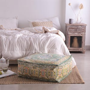 May include: A square, patterned floor pouf with a green, yellow, and pink design. The pouf sits on a woven jute rug in front of a bed with white bedding. A small side table and tea set are also visible.