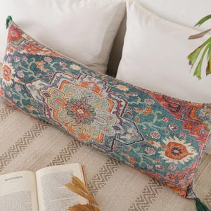 Puede incluir: Un cojín decorativo largo y rectangular con un patrón detallado y colorido en verde azulado, naranja y rojo. El cojín tiene borlas verdes en cada extremo. Descansa sobre una cama con almohadas blancas y una manta beige estampada. Un libro abierto es visible.