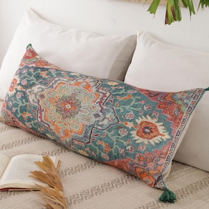 May include: A rectangular decorative pillow with a detailed, colorful pattern in teal, orange, and cream. The pillow has green tassels on each end and is placed on a bed with white pillows.