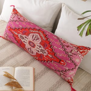 Può includere: Un lungo cuscino lombare rettangolare caratterizzato da un motivo vivace e complesso nei toni del rosa acceso, rosso brillante e crema tenue. Il cuscino è rifinito con piccole nappe rosa su ciascuno dei quattro angoli, aggiungendo un tocco decorativo.