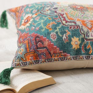 May include: A decorative throw pillow with a teal, orange, and red patterned design. The pillow has a beige backing and green tassels. The pillow is resting on an open book.