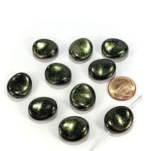 May include: A collection of dark green, oval-shaped beads with a shimmering, glittery effect. The beads are scattered on a white surface, with a penny included for size reference. The beads have a hole for stringing, suitable for jewelry making.