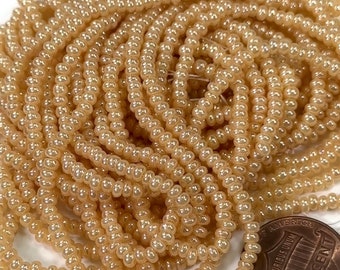 May include: A close-up of a pile of small, round, beige seed beads. The beads have a slight iridescent sheen. A US penny is included for scale.