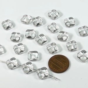 May include: A collection of clear glass square beads with a faceted design. The beads are approximately the size of a US penny.