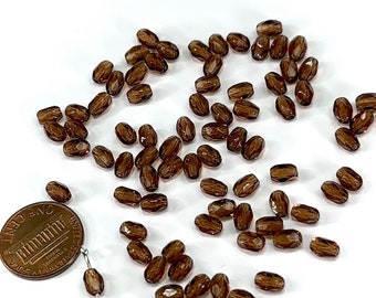 May include: A scattering of brown glass beads with a faceted oval shape. The beads are translucent and have a striated pattern. A US penny is included for scale.
