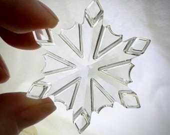 May include: A clear, snowflake-shaped ornament with six points. Each point has a diamond-shaped detail at the end and a series of V-shaped lines. The ornament is held up against a bright background.