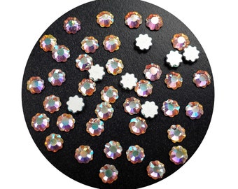 May include: Assortment of iridescent flower-shaped rhinestones on a black background. The rhinestones have a rainbow effect and are scattered across the surface.