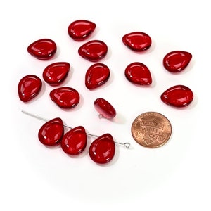 May include: A collection of 14 red glass teardrop beads, some of which are arranged in a line, with a US penny for scale.