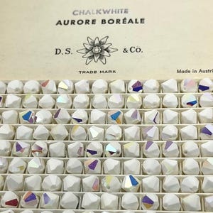 May include: A grid of white, faceted beads with iridescent facets. The box above the beads reads "CHALKWHITE AURORE BORÉALE" with the logo of D.S. & Co. and the words "TRADE MARK". These beads are likely for crafting jewellery.