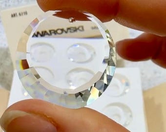 May include: A clear, faceted crystal pendant in a crescent moon shape, held between fingers. The word "SWAROVSKI" is visible on the crystal. The pendant has a small hole at the top for hanging. The background shows a white card with the text "ART 6710".