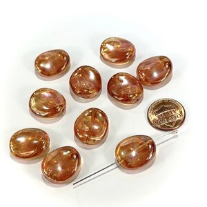 May include: A collection of eleven iridescent, oval-shaped beads in shades of amber and peach. A US penny is included for size reference. A silver-colored beading needle is also present, suggesting the beads are for jewelry making or crafts.