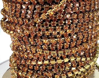 May include: Close-up of a spool of gold-colored rhinestone chain. The chain is made up of small, square rhinestones set in a gold metal base. The rhinestones are a warm, amber color, and the chain is tightly wound.