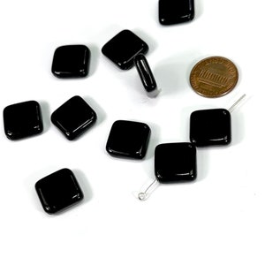May include: A group of black square glass beads scattered on a white surface. A US penny is included for scale.