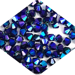 Puede incluir: Una vista de cerca de un montón de cuentas de cristal azul y morado iridiscente. Las cuentas son facetadas y tienen un acabado brillante.