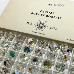 Op de afbeelding: Een doos met heldere, facetgeslepen kristallen kralen met iriserende kleurre reflecties. Op het deksel van de doos staat "CRYSTAL AURORE BORÉALE" en "D.S. & Co." met een bloemenembleem. De kralen zijn in een rasterpatroon gerangschikt.