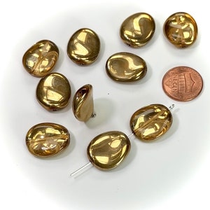 May include: A group of 10 gold-colored glass beads, some are oval shaped and some are more rounded. The beads are arranged on a white surface with a US penny for scale.