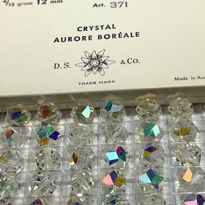 May include: Close-up of a box of clear crystal beads. The beads are faceted and reflect multiple colors. The box has text that reads "CRYSTAL AURORE BORÉALE" and "D.S. & Co.". The beads are 12 mm in size.