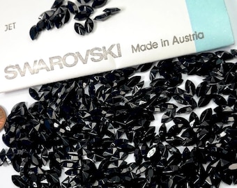 May include: A close-up image of a large quantity of black, faceted, marquise-shaped Swarovski crystals. The crystals are loose and scattered on a white surface. The crystals are unfoiled and made in Austria. The packaging for the crystals is visible in the background.