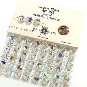 May include: A close-up of a package of 12 mm crystal beads. The beads are clear with an iridescent finish. The package is labeled "1/12 gross 12 mm Art. 346 Aurore Boreale Crystal D.S. & Co. Trade Mark Made in Austria".