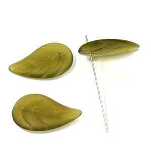 May include: Three olive-green leaf-shaped beads with detailed veining. One bead has a silver wire inserted, suggesting they are components for jewelry or crafts. The beads are set against a white background.