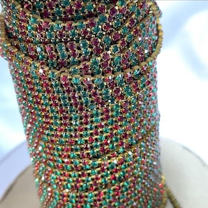 May include: Close-up of a decorative bracelet featuring rows of red and green rhinestones set in a gold-tone metal. The bracelet is tightly coiled, showcasing the intricate design and sparkling details. The overall aesthetic is festive and glamorous.