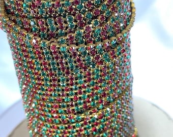 May include: Close-up of a decorative bracelet featuring rows of red and green rhinestones set in a gold-tone metal. The bracelet is tightly coiled, showcasing the intricate design and sparkling details. The overall aesthetic is festive and glamorous.