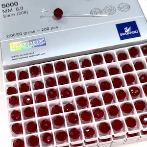 Könnte beinhalten: Eine Schachtel facettierter, tiefroter Swarovski-Kristalle, jeweils etwa 0,8 cm Durchmesser, in einem Raster angeordnet. Die Schachtel ist mit Produktdetails beschriftet, darunter "Siam (208)" und "2/00/00 gross = 288 pcs."