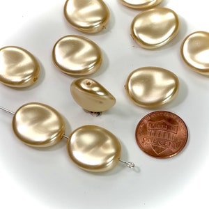 May include: A group of 10 smooth, oval-shaped, cream-colored beads with a pearlescent finish. The beads are arranged in a random pattern on a white surface. A US penny is included for size reference.