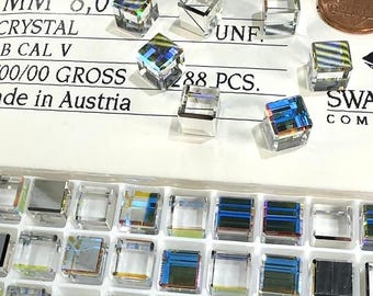 May include: A collection of clear, square-cut crystals with iridescent facets. The crystals are 8.0 mm in size and are labeled with text including "4840", "CRYSTAL", and "288 PCS.". A penny is included for size reference. The crystals are arranged in a tray.