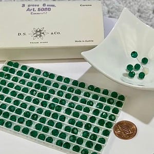 May include: An assortment of emerald green faceted beads, some loose in a white dish and others arranged in a grid. A box with text reading "3 gross 6 mm. Art. 5080 Emerald" and a penny are also present. The box is labeled "Made in Austria."