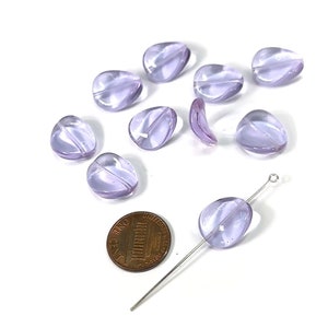 May include: A collection of lavender-colored glass beads in a teardrop shape. One bead is strung on a silver-colored head pin. A U.S. penny is included for size reference. The beads are translucent and have a smooth, glossy finish.